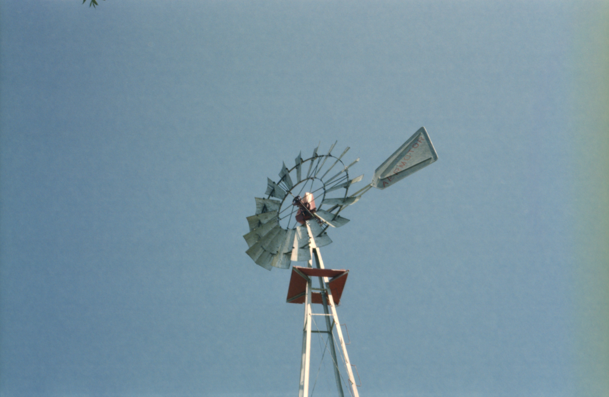 Colour photo of a wind turbine against a blue summer sky. The wind turbine has white blades and a red platform. The lettering AERMOTOR can be seen on the wind vane. The right edge of the photo is slightly yellowish in colour.