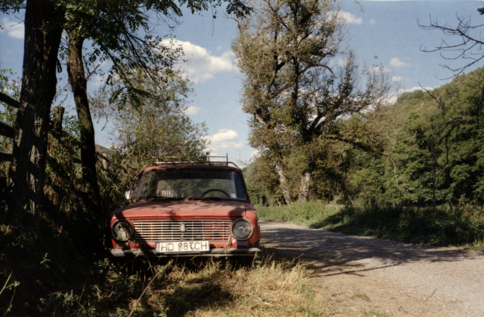 Colour photo of an old red broken-down car on the side of a dirt road. It's a beautiful summer's day with scattered clouds in the sky. There are some trees on both sides of the road, it almost looks as if the car is resting in the shade of the trees.
