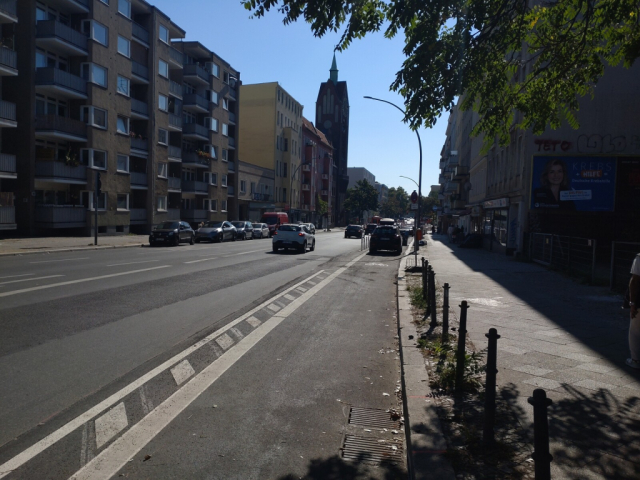 foto des neu eingerichteten radwegs auf der beusselstraße in berlin. der radweg zieht sich vom unteren bildrand in die mitte und ist nach ca. 50m mit autos zugeparkt. der radweg wird begrenzt durch kennzeichnung auf der straße und nach ein paar metern durch poller zur straße. die straße liegt links des weges, befahren von einigen autos. auf der gegenrichtung parken weitere autos. rechts und links der straße stehen typische berliner häuser, im hintergrund links ist ein kirchturm zu sehen. der himmel ist blau, es ist sonnig.