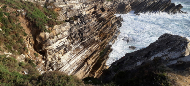 Foto: Um trecho de litoral: a água do mar, azulada e com muitas espumas brancas, encontra uma série de camadas de rocha em tons claros e escuros de marrom, inclinadas a cerca de 45 graus. No canto esquerdo, uma vegetação rala e rasteira cobre parte do solo. ////// Eng: Photo: A stretch of coastline: the blue sea water, with lots of white foam, meets a series of rock layers in light and dark shades of brown, sloping at about 45 degrees. In the left corner, sparse, low vegetation covers part of the ground. ////// CC BY-NC-ND 4.0 2025 Ground Control Foto: Um trecho de litoral: a água do mar, azulada e com muitas espumas brancas, encontra uma série de camadas de rocha em tons claros e escuros de marrom, inclinadas a cerca de 45 graus. No canto esquerdo, uma vegetação rala e rasteira cobre parte do solo. ////// Eng: Photo: A stretch of coastline: the blue sea water, with lots of white foam, meets a series of rock layers in light and dark shades of brown, sloping at about 45 degrees. In the left corner, sparse, low vegetation covers part of the ground. ////// CC BY-NC-ND 4.0 2025 Ground Control