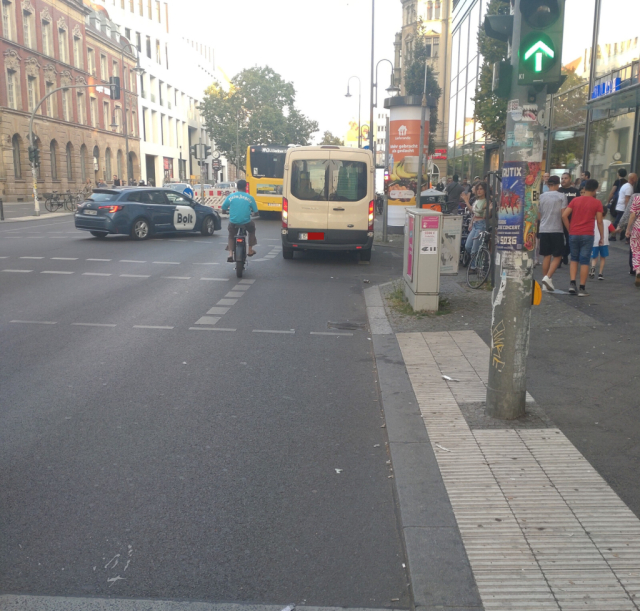 straße in berlin. die straße ist gesäumt von mehrgeschossigen häusern. im vordergrund rechts neben der straße eine ampel mit einem grünen pfeil, rechts daneben ein bürgersteig mit etlichen fußgängern. vom unteren bildrand bis zur bildmitte ein fußgängerübergang von rechts nach links. dahinter beginnt ein radweg, richtung geradeaus an der rechten straßenseite. auf dem radweg steht nach 5 m ein kleintransporter, die warnblinkanlage ist eingeschaltet. der kleintransporter wird links umfahren von einem radfahrer. links neben dem radfahrer ein pkw, der gerade auf die straße einbiegt.