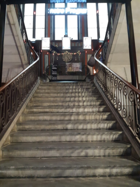 Foto de uma escadaria (no interior do prédio do cinema visto na primeira foto). Há 13 degraus de concreto na escadaria; em ambos os lados, há um corrimão de madeira escura sobre estrutura metálica. Na parede do primeiro andar há um grande relógio e três grandes janelas verticais de vidro. Há também dois letreiros luminosos e três paineis luminosos. A escadaria prossegue, à direita e à esquerda, para um andar superior.<br /><br />============<br /><br />Photo of a staircase (inside the cinema building seen in the first photo). There are 13 concrete steps on the staircase; on both sides, there is a dark wooden handrail on a metal frame. On the wall of the first floor, there is a large clock and three large vertical glass windows. There are also two illuminated signs and three illuminated panels. The staircase continues to the right and left to an upper floor.