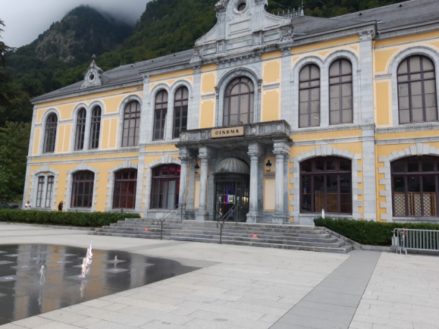 Foto do edifício de um cinema, constituído por térreo e primeiro andar, nas cores amarelo, branco e cinza. A extensão da frente do prédio é de aproximadamente 40 metros. Há uma porta de entrada (com duas colunas de cada lado) e 23 grandes janelas de vidro. Acima da entrada há uma placa onde se lê: "CINEMA". À frente do prédio há uma pequena escadaria de 6 degraus e uma fonte com esguichos d'água. Atrás do pŕedio há montanhas cobertas com vegetação. O céu está muito nublado.<br /><br />================<br /><br />Photo of a cinema building, consisting of a ground floor and first floor, in yellow, white, and gray. The front of the building is approximately 40 meters long. There is an entrance door (with two columns on each side) and 23 large glass windows. Above the entrance there is a sign that reads: “CINEMA.” In front of the building there is a small staircase with 6 steps and a fountain with water jets. Behind the building there are mountains covered with vegetation. The sky is very cloudy.
