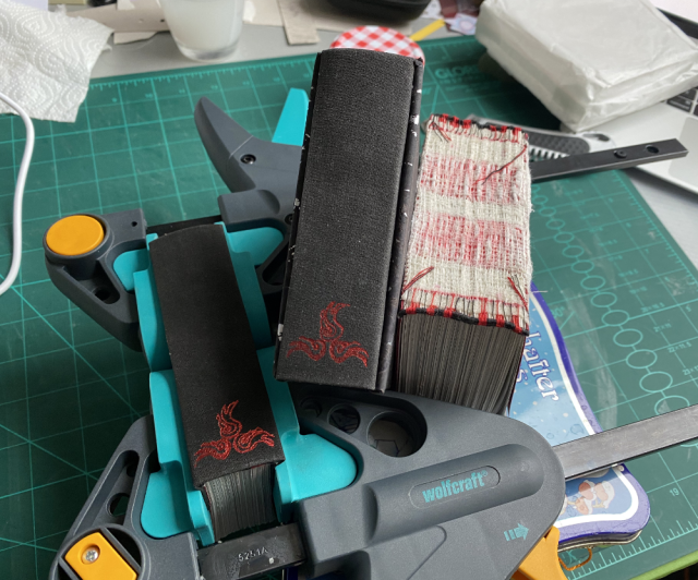 Photo shows three books in various states of finished. One book is clamped with two C-clamps as the spine dries. The other is finished and the third and thickest is just a textbloock. Photo shows three books in various states of finished. One book is clamped with two C-clamps as the spine dries. The other is finished and the third and thickest is just a textbloock.