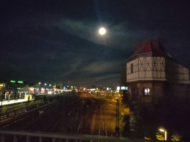 unscharfes nachtfoto des mondes. von einer erhöhung fotographiert, unten verlaufen schienen in richtung bildmitte. links, parallel dazu die lichter eines s-bahnhofes. am horizont in der bildmitte weitere verschwommene lichter in rot, gelb und weiß. rechts ein altertümlich wirkendes gebäude der bahn in hellem fachwerk mit rotem dach. der mond in der oberen bildmitte wird von einigen schleierwolken bedeckt