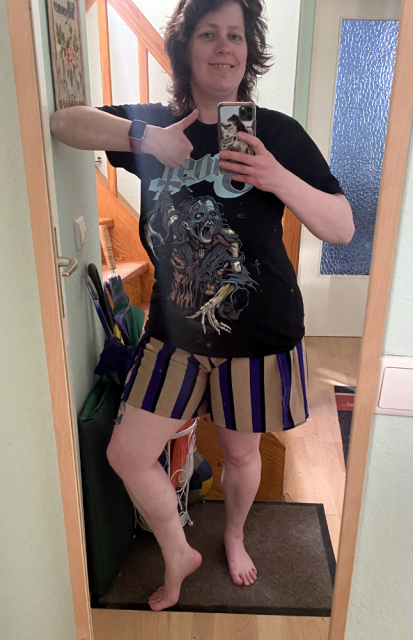 A woman taking a selfie in the mirror. She's giving a thumbs up and is wearing a Ghost shirt and trousers ending abov the knees, light brown with thick blue, vertical stripes. A woman taking a selfie in the mirror. She's giving a thumbs up and is wearing a Ghost shirt and trousers ending abov the knees, light brown with thick blue, vertical stripes.