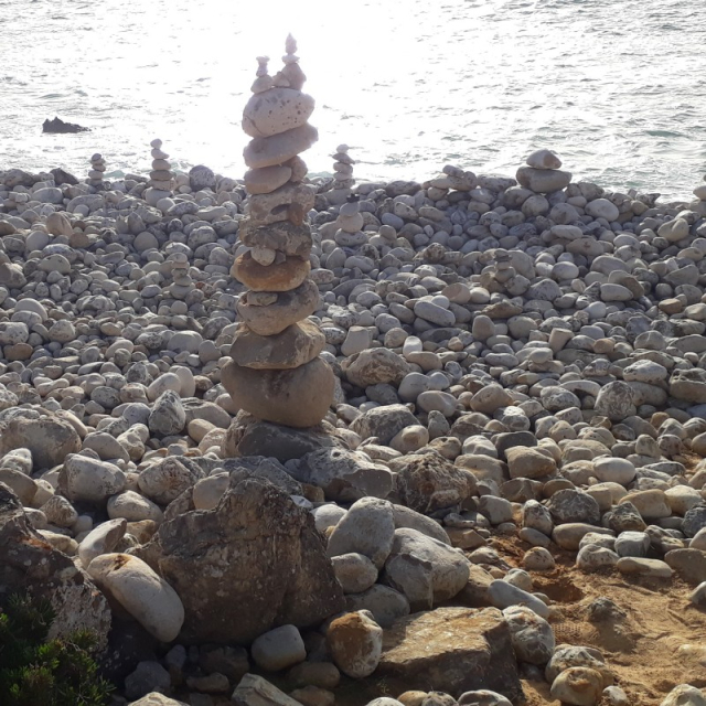 Foto: Um trecho de uma praia, quase inteiramente coberto por pedras arredondadas no chão. Há uma torre de pedras empilhadas (próximo ao centro da imagem), com cerca de um metro de altura, formada por cerca de 20 pedras de tamanhos diferentes. Alguns metros atrás, há outras três torres bem menores, formadas por cinco ou seis pequenas pedras. A luz do sol é refletida intensamente na água do mar. ////// Eng: Photo: A stretch of beach, almost entirely covered with rounded stones on the ground. There is a tower of stacked stones (near the center of the image), about one meter high, formed by about 20 stones of different sizes. A few meters behind, there are three other much smaller towers, formed by five or six small stones. The sunlight is reflected intensely on the sea water. ////// CC BY-NC-ND 4.0 2025 Ground Control Foto: Um trecho de uma praia, quase inteiramente coberto por pedras arredondadas no chão. Há uma torre de pedras empilhadas (próximo ao centro da imagem), com cerca de um metro de altura, formada por cerca de 20 pedras de tamanhos diferentes. Alguns metros atrás, há outras três torres bem menores, formadas por cinco ou seis pequenas pedras. A luz do sol é refletida intensamente na água do mar. ////// Eng: Photo: A stretch of beach, almost entirely covered with rounded stones on the ground. There is a tower of stacked stones (near the center of the image), about one meter high, formed by about 20 stones of different sizes. A few meters behind, there are three other much smaller towers, formed by five or six small stones. The sunlight is reflected intensely on the sea water. ////// CC BY-NC-ND 4.0 2025 Ground Control