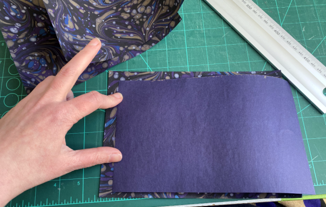 A piece of marbled paper lies on the cutting mat, a smaller piece is held over it, purple backside up. A piece of marbled paper lies on the cutting mat, a smaller piece is held over it, purple backside up.