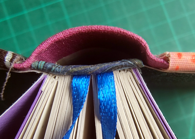 A close up at the opened book. We look into the spine and at the glued on paper endbands. The two blue ribbons grow from beneath the endbands. A close up at the opened book. We look into the spine and at the glued on paper endbands. The two blue ribbons grow from beneath the endbands.